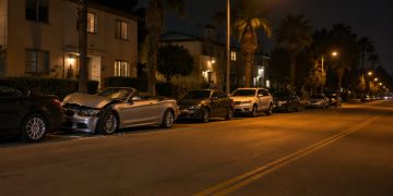 Damaged silver convertible pressed against a row of parked cars on a residential street of apartment buildings and palm trees in University Park at night.