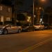 Damaged silver convertible pressed against a row of parked cars on a residential street of apartment buildings and palm trees in University Park at night.