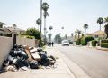 Daytime residential street in the Fairfax District of Los Angeles with piled trash bags and debris beside a fence in the foreground and homes blurred behind.