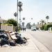 Daytime residential street in the Fairfax District of Los Angeles with piled trash bags and debris beside a fence in the foreground and homes blurred behind.