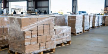 Daytime view of pallets of boxed shoes and goods inside a Southern California warehouse, with a blurred loading dock in the background.