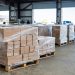Daytime view of pallets of boxed shoes and goods inside a Southern California warehouse, with a blurred loading dock in the background.