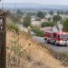 Dry hillside brush at the edge of a West Covina neighborhood in daylight, with a fire engine blurred on the road in the background.