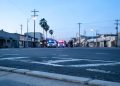 Early-morning view of a Santa Ana street with empty lanes and a curb in the foreground and emergency vehicle lights blurred in the distance.