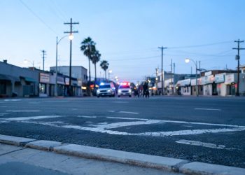 Early-morning view of a Santa Ana street with empty lanes and a curb in the foreground and emergency vehicle lights blurred in the distance.