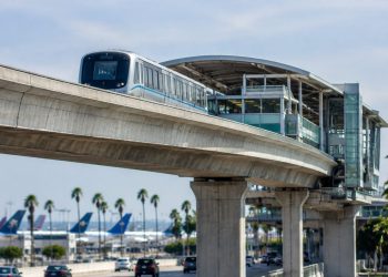 Elevated concrete guideway and station beside an airport access road at Los Angeles International Airport in daylight, palm trees blurred behind.