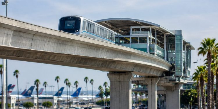 Elevated concrete guideway and station beside an airport access road at Los Angeles International Airport in daylight, palm trees blurred behind.