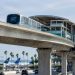 Elevated concrete guideway and station beside an airport access road at Los Angeles International Airport in daylight, palm trees blurred behind.