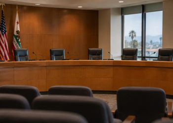 Empty city council dais with a curved wooden desk, microphones and high-backed chairs in a municipal chamber, illustrating West Hollywood's multi-partner ordinance.