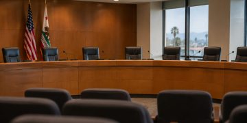 Empty city council dais with a curved wooden desk, microphones and high-backed chairs in a municipal chamber, illustrating West Hollywood's multi-partner ordinance.