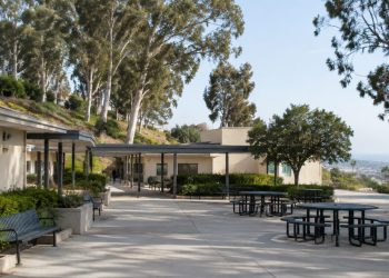 Empty courtyard of a hillside school campus in Brentwood with low stucco buildings, a covered walkway and eucalyptus trees in afternoon daylight.