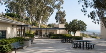 Empty courtyard of a hillside school campus in Brentwood with low stucco buildings, a covered walkway and eucalyptus trees in afternoon daylight.
