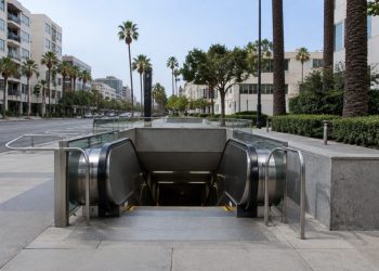 Empty escalator descending into a subway station entrance on Wilshire Boulevard in Los Angeles, with palm trees and mid-rise buildings in daylight.