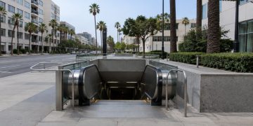 Empty escalator descending into a subway station entrance on Wilshire Boulevard in Los Angeles, with palm trees and mid-rise buildings in daylight.