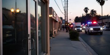 Evening street in Florence-Graham with a closed business entrance in the foreground and emergency vehicle lights blurred far in the background.