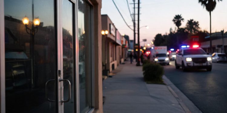 Evening street in Florence-Graham with a closed business entrance in the foreground and emergency vehicle lights blurred far in the background.