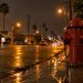 Fire hydrant at a South Los Angeles street corner at night with water pooled on wet asphalt under street lamps, low-rise buildings softly blurred behind.