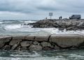 Fractured concrete bulkhead in sharp focus, with churning Pacific swells and rocky shore softly blurred behind, showing coastal storm damage, Southern California.