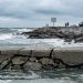 Fractured concrete bulkhead in sharp focus, with churning Pacific swells and rocky shore softly blurred behind, showing coastal storm damage, Southern California.