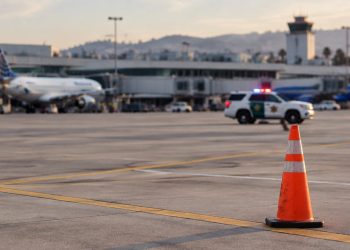 Late-afternoon view of an airport apron at John Wayne Airport with a traffic cone in the foreground and a patrol SUV blurred in the distance.