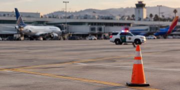 Late-afternoon view of an airport apron at John Wayne Airport with a traffic cone in the foreground and a patrol SUV blurred in the distance.