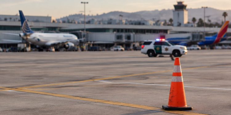 Late-afternoon view of an airport apron at John Wayne Airport with a traffic cone in the foreground and a patrol SUV blurred in the distance.