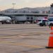 Late-afternoon view of an airport apron at John Wayne Airport with a traffic cone in the foreground and a patrol SUV blurred in the distance.