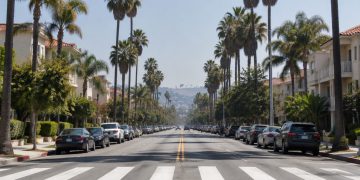 Long-lens view down Fountain Avenue in West Hollywood with parked cars along the curbs, a painted crosswalk, palm trees and low-rise apartment buildings in daylight.