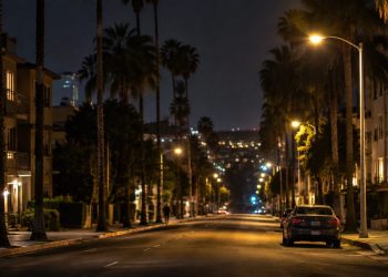 Long-lens view down North Fairfax Avenue in West Hollywood at night, with low-rise apartment buildings, palm trees and a parked sedan under street lamps.