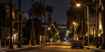 Long-lens view down North Fairfax Avenue in West Hollywood at night, with low-rise apartment buildings, palm trees and a parked sedan under street lamps.