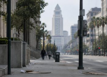 Long-lens view down an emptied Spring Street in downtown Los Angeles toward City Hall in afternoon daylight, with scattered litter on the sidewalk.