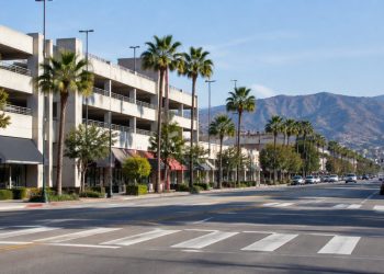 Long-lens view of a commercial street in downtown Burbank with a multilevel parking structure, low-rise storefronts and palm trees in afternoon daylight.