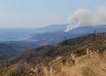 Long-lens view of a dry chaparral canyon in the Santa Monica Mountains near Malibu with utility lines on the ridge and a smoke plume over distant hills.