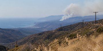 Long-lens view of a dry chaparral canyon in the Santa Monica Mountains near Malibu with utility lines on the ridge and a smoke plume over distant hills.