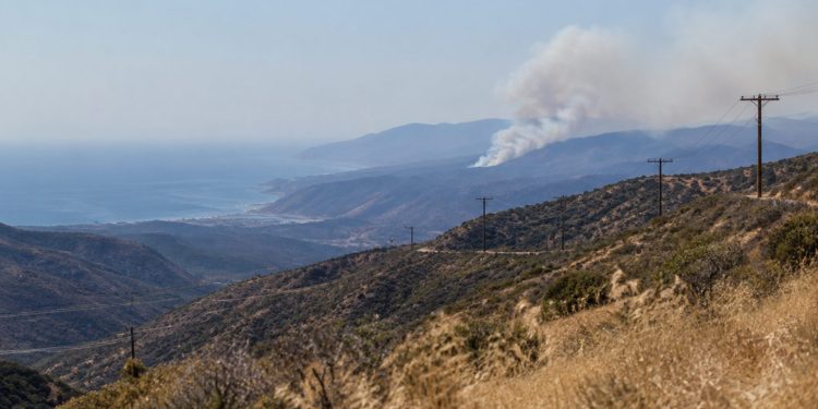 Long-lens view of a dry chaparral canyon in the Santa Monica Mountains near Malibu with utility lines on the ridge and a smoke plume over distant hills.