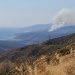 Long-lens view of a dry chaparral canyon in the Santa Monica Mountains near Malibu with utility lines on the ridge and a smoke plume over distant hills.