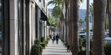 Long-lens view of a palm-lined commercial street of low-rise shops in West Hollywood in neutral daylight, with pedestrians softly blurred.