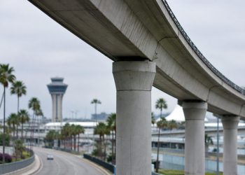 Long-lens view of an empty elevated concrete guideway curving above an access road at Los Angeles International Airport in neutral daylight.