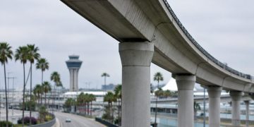 Long-lens view of an empty elevated concrete guideway curving above an access road at Los Angeles International Airport in neutral daylight.