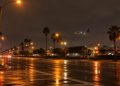 Long-lens view of an empty multi-lane boulevard intersection at night near Los Angeles International Airport, with an airliner descending in the distance.