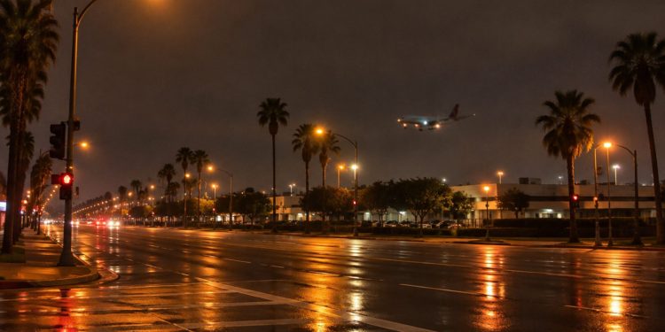 Long-lens view of an empty multi-lane boulevard intersection at night near Los Angeles International Airport, with an airliner descending in the distance.