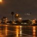 Long-lens view of an empty multi-lane boulevard intersection at night near Los Angeles International Airport, with an airliner descending in the distance.