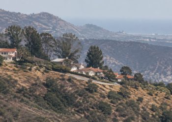 Long-lens view of dry chaparral slopes and hillside homes along a ridge in the Santa Monica Mountains of Los Angeles in neutral daylight.