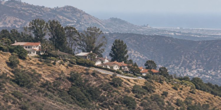 Long-lens view of dry chaparral slopes and hillside homes along a ridge in the Santa Monica Mountains of Los Angeles in neutral daylight.