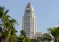Los Angeles City Hall tower in neutral daylight, seen from a low angle above softly blurred palm trees, illustrating the city and county homelessness dispute.