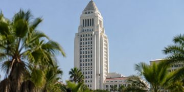 Los Angeles City Hall tower in neutral daylight, seen from a low angle above softly blurred palm trees, illustrating the city and county homelessness dispute.