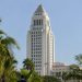 Los Angeles City Hall tower in neutral daylight, seen from a low angle above softly blurred palm trees, illustrating the city and county homelessness dispute.