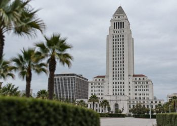 Los Angeles City Hall tower in neutral daylight, sharp above softly blurred palm trees, illustrating the debate over 2028 Olympics finances.