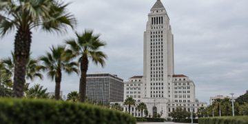 Los Angeles City Hall tower in neutral daylight, sharp above softly blurred palm trees, illustrating the debate over 2028 Olympics finances.