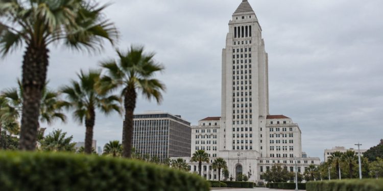 Los Angeles City Hall tower in neutral daylight, sharp above softly blurred palm trees, illustrating the debate over 2028 Olympics finances.